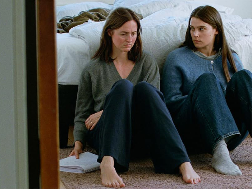 Two frowning women sit on a rug with their backs against an unmade bed; one woman holds a spiral notebook